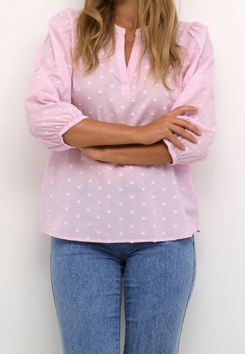 Femme aux longs cheveux portant un chemisier rose clair texturé à manches trois-quarts et un jean bleu, bras croisés au niveau de la taille.