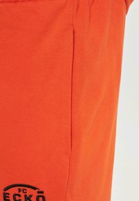 Orange cotton shorts with a smooth texture, featuring side pockets and black embroidered logo detail at the lower left.