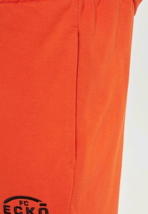 Orange cotton shorts with a smooth texture, featuring side pockets and black embroidered logo detail at the lower left.