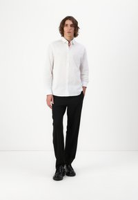 Cămașă albă cu mânecă lungă, cu nasturi, dintr-un material texturat, combinată cu pantaloni negri și pantofi negri; modelul stă în fața unui fundal simplu.