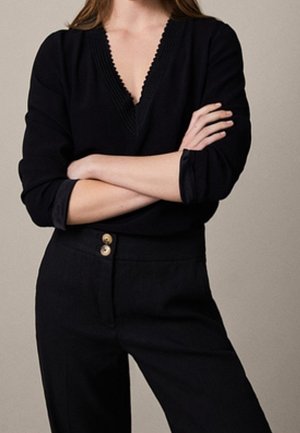 Donna che indossa una maglia nera a maniche lunghe con scollo a V e bordi smerlati, e pantaloni neri con due bottoni, con le braccia incrociate sul petto.