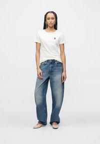 White short-sleeve t-shirt with a small black heart logo, paired with loose-fitting blue denim jeans, on a neutral background.