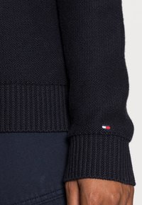 Morski sweter dzianinowy z ribbami na mankietach, z małą poziomą naszywką w trzech kolorach. Gładka faktura, klasyczny okrągły dekolt.