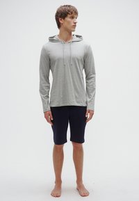 Sweatshirt cinzento claro de manga comprida com capuz e cordões, combinado com calções azul-marinho até ao joelho. Modelo está descalço, posando contra um fundo branco.