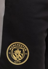 Στρογγυλό λογότυπο της Manchester City σε χρυσό πάνω σε μαύρο ύφασμα, που δείχνει ένα πλοίο, μια ασπίδα και ένα τριαντάφυλλο με το έτος 1894.