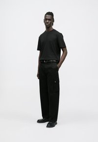 Μαύρο βαμβακερό t-shirt με κοντά μανίκια, συνδυασμένο με άνετο μαύρο παντελόνι τύπου καρό με πλαϊνές τσέπες, φορεμένο με μαύρα παπούτσια.