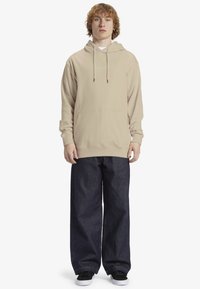 Felpa beige con tasca frontale e cordino regolabile. Indossata con pantaloni in denim scuri e larghi e sneakers nere.