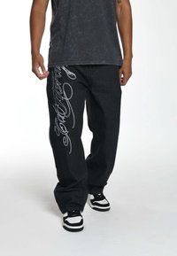 Zwarte spijkerbroek heeft een losse pasvorm met witte cursieve borduursels langs het linkerbeen. Gecombineerd met een donker T-shirt en zwart-witte sneakers.