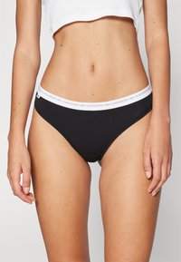 Slip en bikini noir avec une ceinture logo Lacoste blanche, fabriqué dans un tissu lisse et extensible, présentant une coupe classique taille basse.