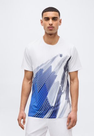 Fiatal férfi fehér pólóban, amelyen absztrakt kék és fekete átlós csíkok találhatók, egy egyszínű világos háttér előtt áll.