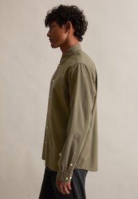 Koszula z długim rękawem na guziki w oliwkowym zielonym kolorze, wykonana z bawełnianej tkaniny, z klasycznym kołnierzem, zaokrąglonym dołem i białymi guzikami. Gładka faktura z minimalną ilością detali.