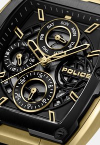 Zwarte en gouden watch heeft een skeletonized wijzerplaat, grote cirkelvormige sub-wijzers en gemarkeerde dagindicatoren, met het "POLICE"-logo prominent weergegeven.