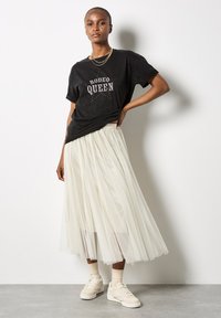 Schwarzes T-Shirt mit "RODEO QUEEN" Grafik, kombiniert mit einem hellcremefarbigen Tüllrock. Das Model trägt beiges Sneaker und geschichtete Halsketten.