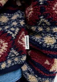 Gevoerde fleece stof in marineblauw, bordeauxrood en crème met een geometrisch patroon. Bevat een etiket met de tekst "ORIGINALS" aan de zijkant.