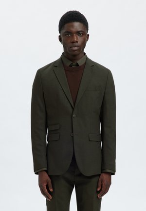 Jeune homme portant un costume vert foncé, un pull marron et une chemise vert foncé sur un fond clair uni, debout et face à l'avant.