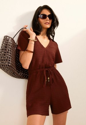 Mujer que lleva un mono burdeos texturizado con cordón, gafas de sol oscuras, pulseras y lleva un gran bolso grande tejido marrón al hombro.