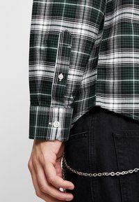 Vihreä-mustaruudullinen paita valkoisilla yksityiskohdilla, jossa on nappikiinnitteinen kalvosin ja rintatasku. Käsi näkyvissä pitelemässä hopeaketjua.