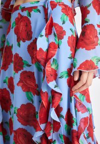 Maxi skirt in light blue fabric featuring large red roses with green leaves. Ruffled accents along the edges. Smooth texture.