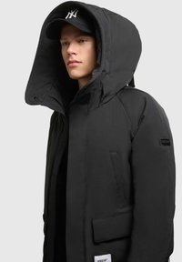 Juodasis izoliuotas parka tipo švarkas su dideliu gobtuvu, užtrauktuku priekyje ir šoninėmis kišenėmis. Yra tekstūruotas paviršius ir prekės ženklas ant rankovės.