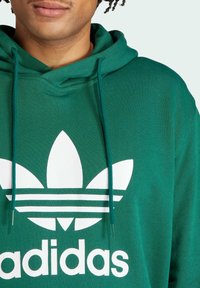 Zelená mikina vyrobená z textilu, s veľkým bielym logom Adidas so štyrlistovou kvetinou a tromi vodorovnými pruhmi. Viditeľné šnúrky.