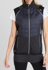 Femme portant un gilet matelassé noir sans manches sur un t-shirt blanc à manches courtes, associé à un legging de sport noir.