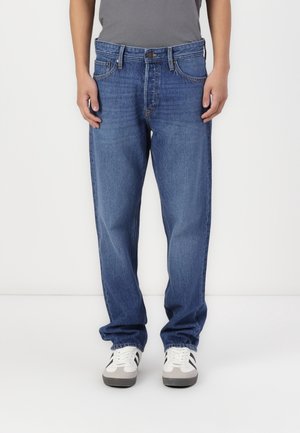 Person trägt lockere blaue Jeans und weiße Sneakers mit schwarzen Streifen, steht vor einem weißen Hintergrund.