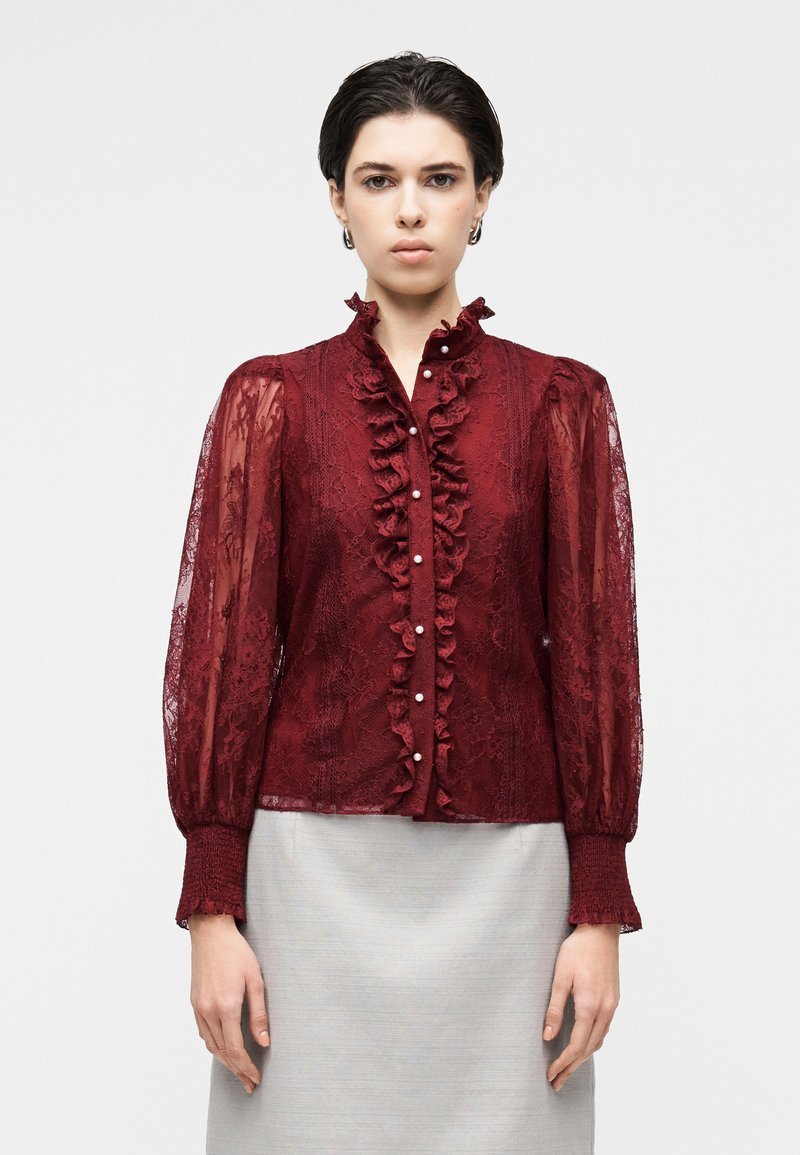 Burgundská čipkovaná blúzka s volánovým golierom, perlovými gombíkmi a priesvitnými dlhými rukávmi s elastickými manžetami, v kombinácii so svetlosivou sukňou.