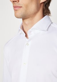 Camicia bianca a maniche lunghe con colletto a punta, caratterizzata da una texture liscia e un leggero luccichio, mostrata da un angolo con bottoni visibili.