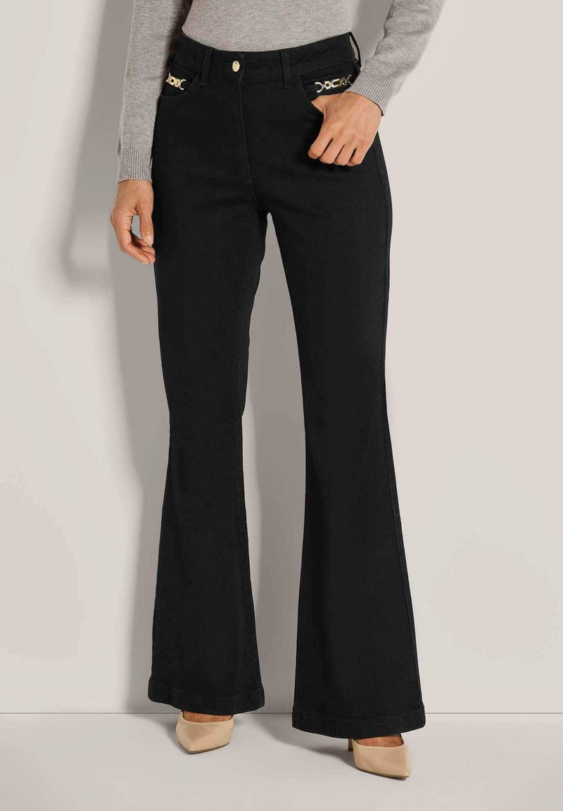 Vrouw in zwarte flared broeken met gouden kettingdetails op de zakken, beige puntige hakken en een grijze langstijdige top, staande tegen een vlakke muur.