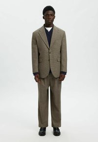 Brūns teksturēts bleizeris ar atbilstošām biksēm, apvienots ar tumši zilu adītu džemperi, pabeigts ar melniem apaviem. Vienkāršs, klasiskos dizains.