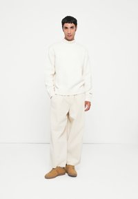 Suéter de cuello alto crema, pantalones beige de corte holgado y zapatillas color marrón claro. El suéter tiene puños de canalé; los pantalones cuentan con una cintura con cordón.