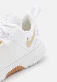 Detailní záběr na bílé tenisky Nike se zlatým logem, síťovaným svrškem, bílými tkaničkami a béžovou gumovou podrážkou na bílém pozadí.