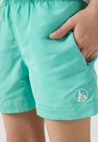 Turquoise zwembroek met een trekkoord in de taille, zijzakken en een wit logo-opgenaaid op de onderste zoom. Gladde, lichte stof.