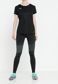Maglietta sportiva nera con accenti e logo bianchi, abbinata a leggings neri con motivi a punti. Calzature includono scarpe da calcio verdi e argento.