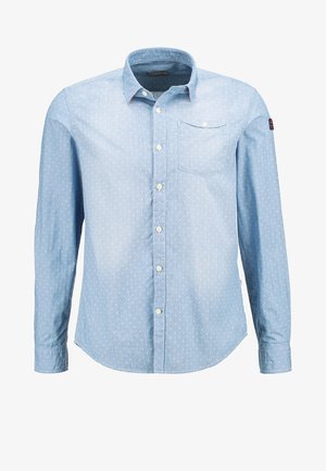 Mand iført lyseblå skjorte med knapper og op rullede ærmer, slidte blå jeans og grå lave sneakers i en afslappet positur.