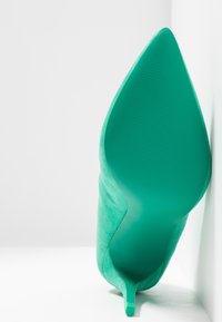 Chaussure à talons hauts en daim vert avec un bout pointu, une surface texturée et une semelle lisse et élégante. Le talon présente un design stilettos.