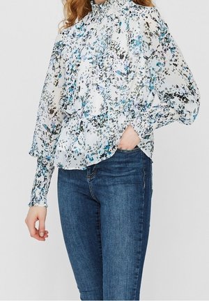 Vrouw draagt een doorschijnende witte blouse met blauw- en zwartgekleurde bloemenpatronen en een hoge kraag, gecombineerd met donkerblauwe strakke jeans.