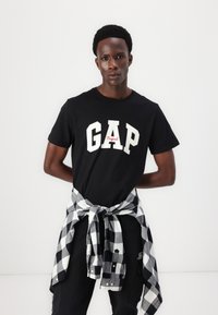 Black t-shirt with "GAP" in bold white letters and red script underneath. Black and white checkered shirt tied around the waist.