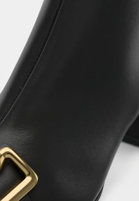 Botte de cheville en cuir noir avec un accent de boucle carrée en or. La surface est lisse avec un design élégant et ajusté.