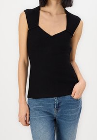 Schwarzes, geripptes, ärmelloses Top mit V-Ausschnitt und strukturierten Schulterdetails, kombiniert mit blauen Jeans und einer figurbetonten Silhouette.