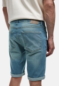 Hombre vistiendo pantalones cortos de mezclilla azul claro arremangados y una camisa blanca, mostrado desde atrás y desde la cintura hacia abajo.