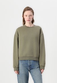 Olijfgroen sweatshirt, comfortabele pasvorm, ronde hals, verlaagde schouders, ribgebreide boorden en onderrand, gedragen met lichtblauwe jeans.