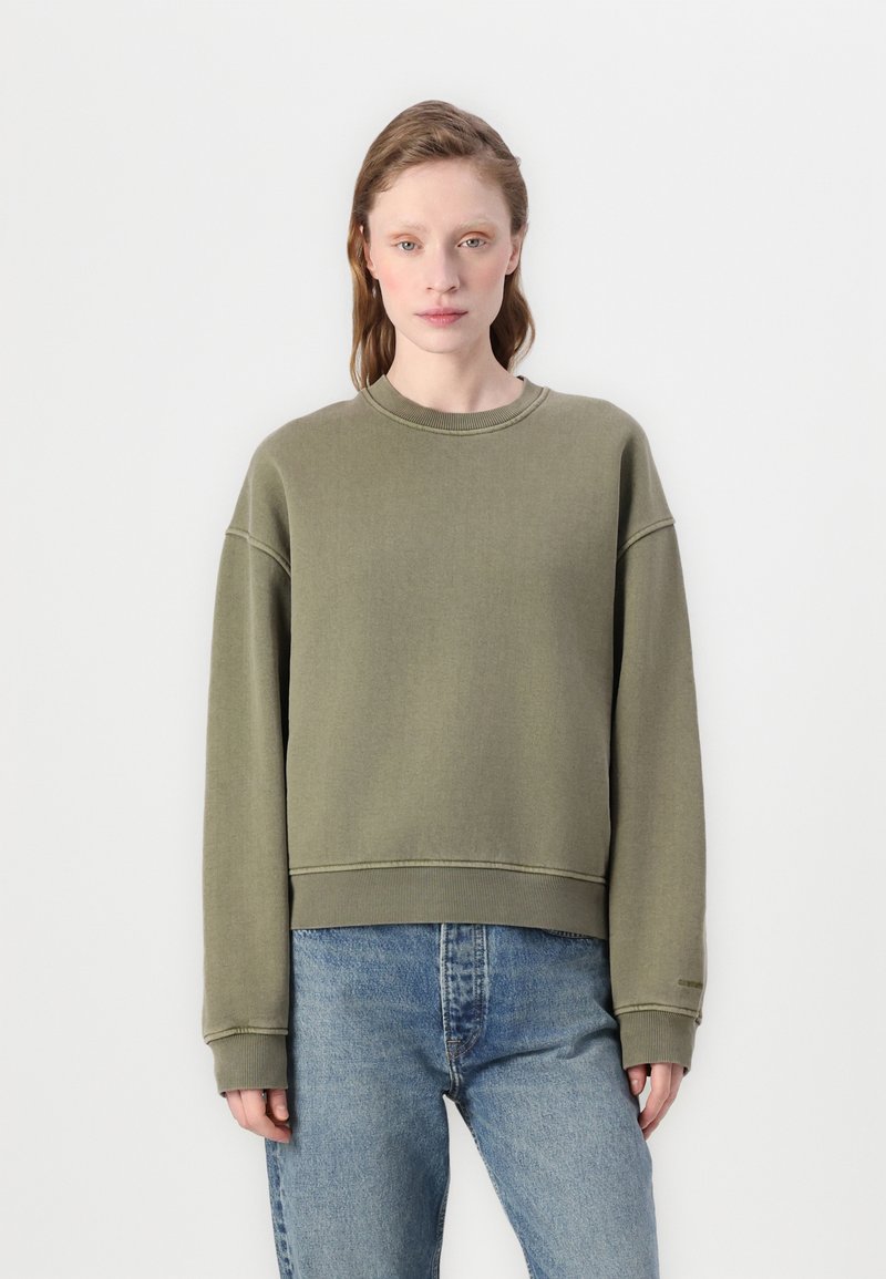Olijfgroen sweatshirt, comfortabele pasvorm, ronde hals, verlaagde schouders, ribgebreide boorden en onderrand, gedragen met lichtblauwe jeans.