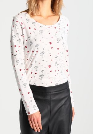 Vrouw draagt een wit top met lange mouwen met rode lippen, zwarte rozen, hartjes en de tekst "love", gecombineerd met een zwarte leren broek met hoge taille.
