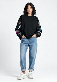 Čierna mikina so grafickými vzormi v ružovej, červenej a modrej farbe na rukávoch, v kombinácii s svetlo modrými rolovanými džínami a bielymi teniskami.