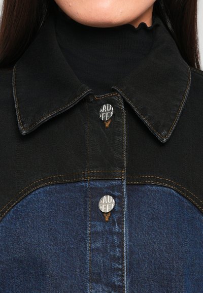 Crna i plava traper jakna s ovratnikom, koja ima metalne dugmiće, kontrastne teksture i vidljive detalje šivanja.