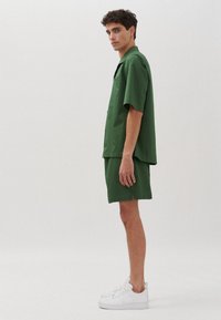 Camicetta verde a maniche corte abbinata a pantaloni corti coordinati, entrambi realizzati in tessuto liscio, presenta una vestibilità comoda e un design minimalista. Indossata con sneakers bianche.