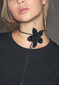 Svart läderhalsband med en blommönster bestående av sex kronblad och metalldetaljer. Bärs runt halsen med ett långt, justerbart band.