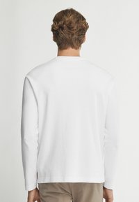 Langärmliges weißes T-Shirt aus Baumwolle, Rundhals, normale Passform, schlichtes Design, mit einem kleinen Logotag am Saum auf der linken Seite.