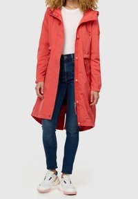 Kobieta w koralowej długiej kurtce, białej bluzce, ciemnoniebieskich przylegających dżinsach i białych sneakersach, stojąca na jednolitym tle.